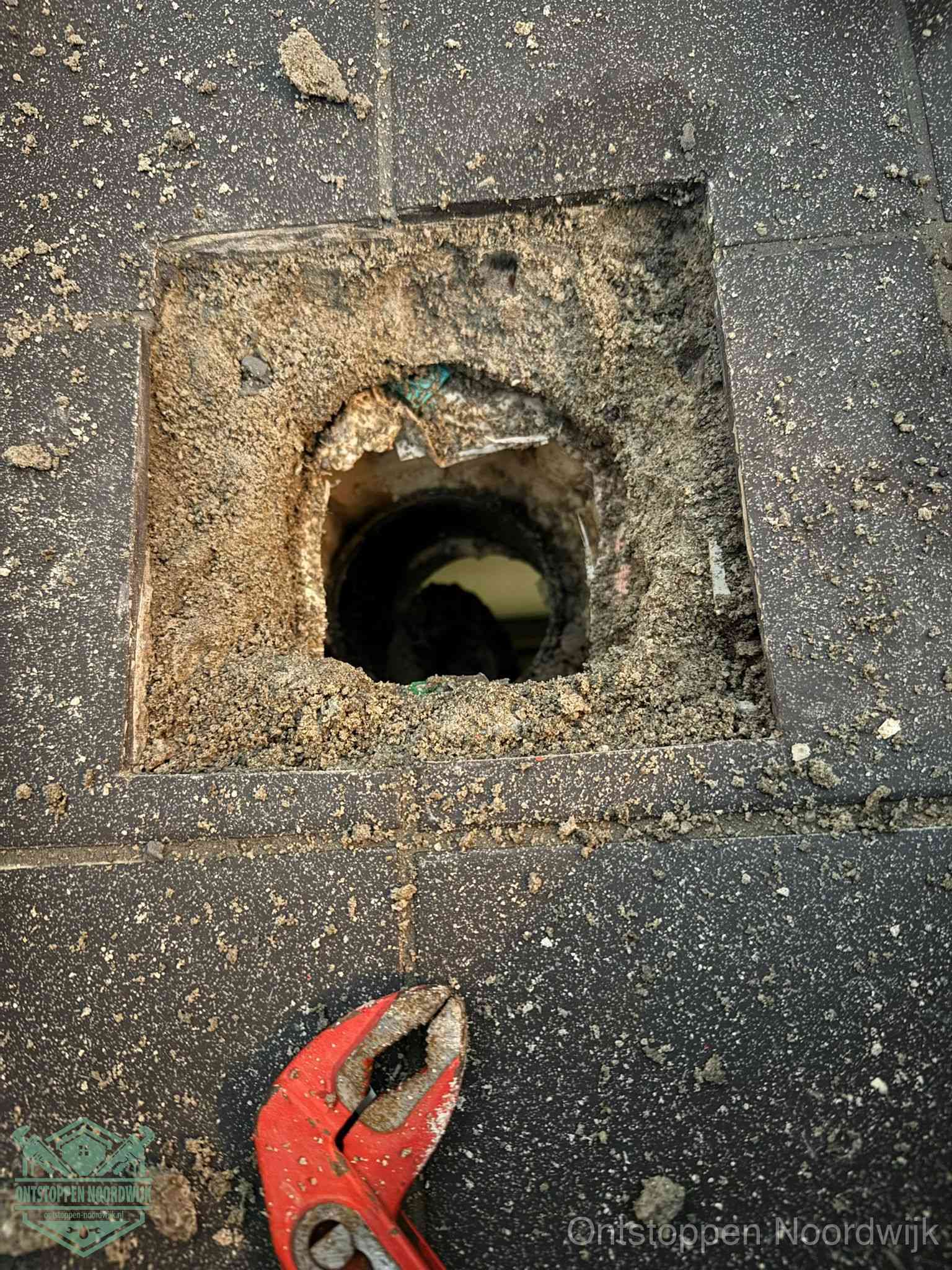Een geopend rioolputje in een betontegelvloer met losgemaakt rooster en een zichtbare ronde afvoerbuis, het achterhalen waar de afvoerverstopping zit in Noordwijk