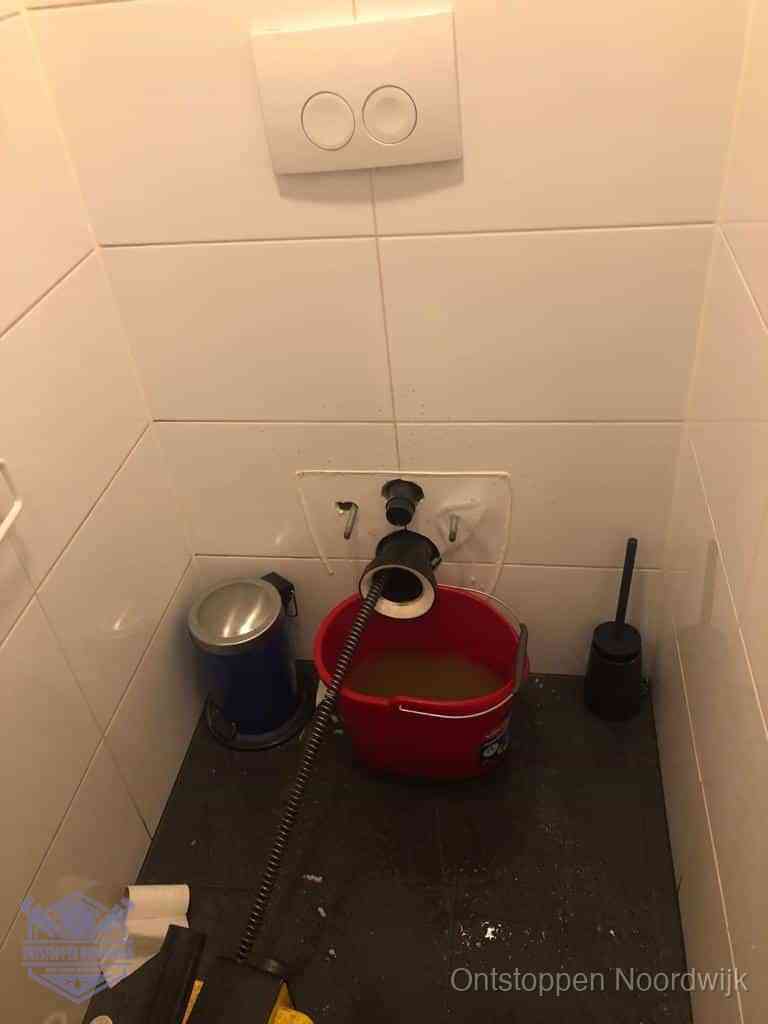 Een ontstopte wand-wc met verwijderde toiletpot waar een ontstoppingsveer in de wanduitgang verdwijnt, het achterhalen waar de verstopping zit in Noordwijk