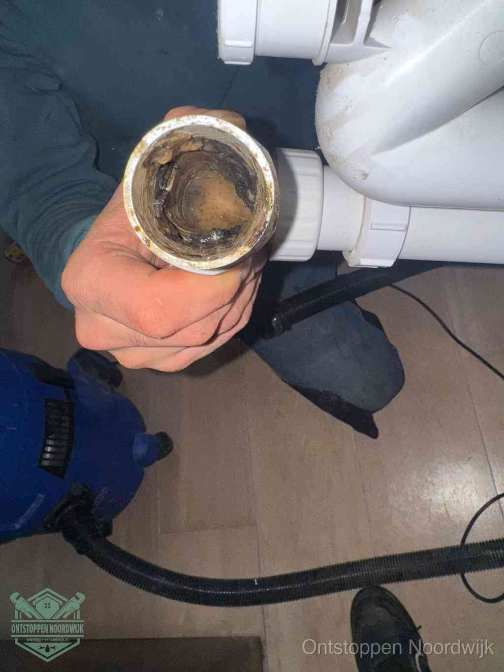 Een hand houdt een losgekoppeld PVC-afvoerbuisstuk vast dat van binnen volledig is dichtgeslibd met vet, vuil en aanslag, de meest voorkomende oorzaak van een verstopte afvoer in Noordwijk