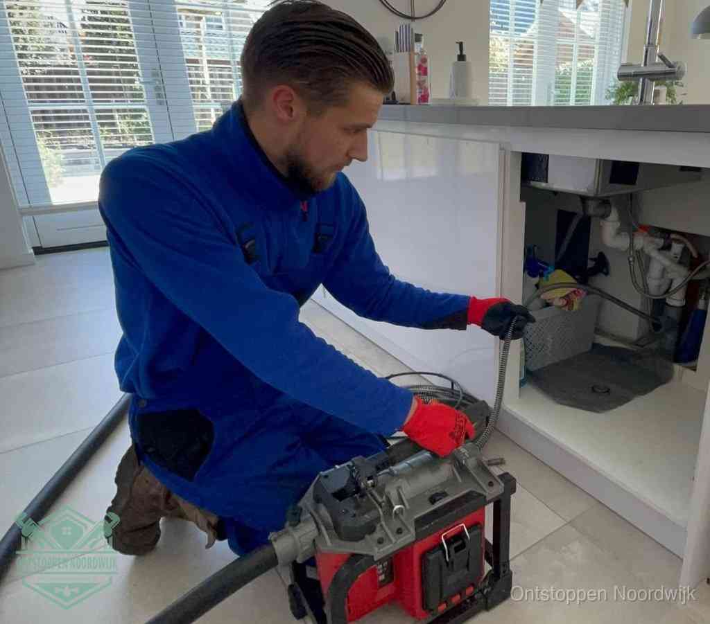 Een monteur met rode handschoenen knielt in een keuken en voert een elektrische ontstoppingsveer in tijdens het professioneel afvoer ontstoppen in Noordwijk